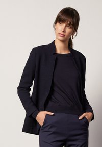 Teksturowana, ciemnogranatowa kurtka z otwartym przodem i bez guzików, połączona z dopasowaną granatową bluzką, noszona z granatowymi spodniami o wysokim stanie.