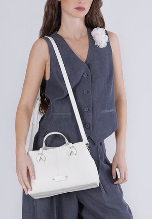 Vrouw met een mouwloos donkergrijs vest met een witte bloemenpin, draagt een witte handtas met schouderriem en handvaten.