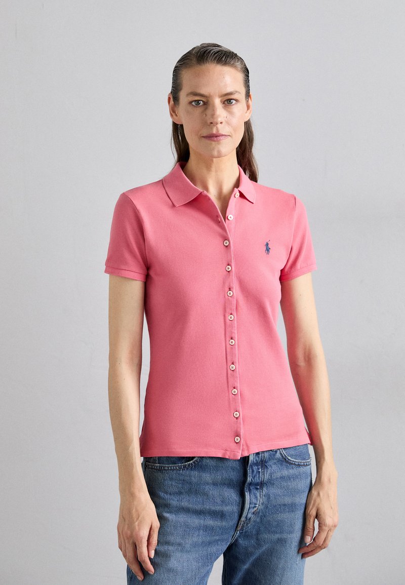 Rosa Poloshirt mit kurzen Ärmeln, Knopfleiste und kleinem blauen Logo auf der linken Brust. Getragen mit hellblauen Denim-Jeans.