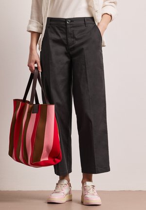 Personne portant un pantalon noir à jambes larges, des baskets rose clair et blanches, tenant un grand sac cabas rayé rouge, rose et brun dans la main gauche.