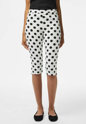 Donna con pantaloni bianchi al ginocchio con pois neri, top nero senza maniche e ballerine nere, in piedi davanti a uno sfondo semplice.