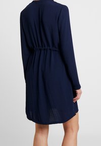 Robe courte bleu marine à manches longues, taille froncée, tissu texturé, et col rond. Longueur au-dessus des genoux, coupe ample.