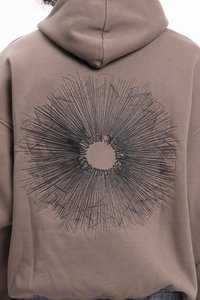 Világosbarna anyagú kapucnis pulóver, amelynek hátulján hangsúlyos fekete grafikai minta látható, ami sugárzó vonalakra hasonlít.