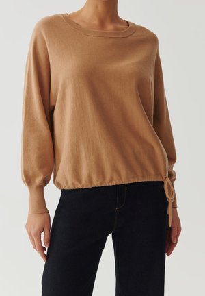 Femme portant un pull ample beige à manches longues avec un cordon de serrage à l’ourlet, associé à un jean bleu foncé sur un fond uni.