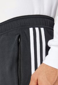Black jogger pants with a soft texture; features white three-stripe design and a zippered pocket on the side. Elastic waistband visible.