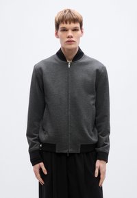 Γκρι μπουφάν τύπου βομβιστή με υφή, μαύρο ριμπ κολάρο και μανσέτες. Μπροστινό φερμουάρ κλεισίματος και πλαϊνές τσέπες. Απλός, αδιάκοπος σχεδι ασμός.