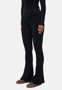 Zwarte ribbelachtige uitlopende broek met een hoge tailleband, kenmerkende naaddetails, zachte textuur en gecombineerd met bruine instappers.