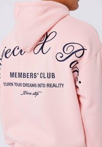 Rosa hoodie met capuchon en donkerblauwe geborduurde tekst, met zinnen zoals "LIDMATSCHAP" en "VERANDER JE DROOMEN IN REALITEIT." Gladde textuur.