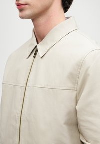 Šviesiai smėlio spalvos bomberio striukė su apykakle, turinti priekinį užtrauktuką ir minimalų siūlių apdailą. Lygi tekstūra.