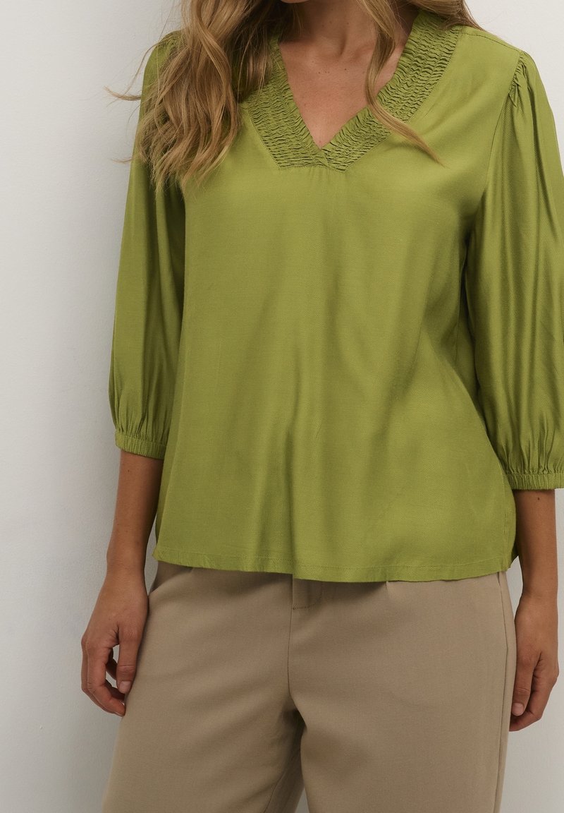 Femme portant un chemisier vert clair à col en V avec manches 3/4 et un pantalon beige, debout devant un fond uni clair.