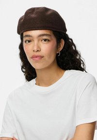 Boina de fieltro marrón con forma redondeada, que presenta una textura suave. Se lleva con una camiseta blanca sencilla, destacando un diseño minimalista.