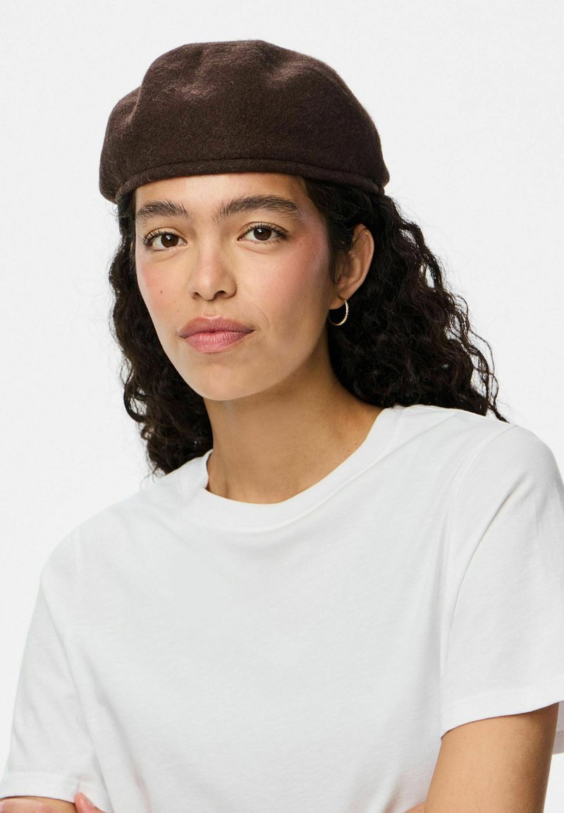Boina de fieltro marrón con forma redondeada, que presenta una textura suave. Se lleva con una camiseta blanca sencilla, destacando un diseño minimalista.