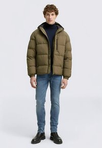 Olivengrøn puffjakke med lynlåslukning og hætte, med quiltet tekstur. Båret over en mørk rullekrave og kombineret med blå jeans.