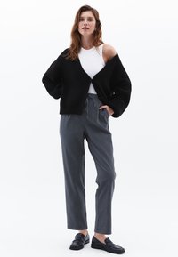 Zwarte oversized cardigan met een V-hals en lange mouwen, gecombineerd met grijze gepaste broek en zwarte platte loafers. Soepele texturen overal.