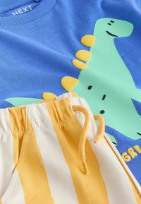 Blauwe katoenen T-shirt met een groene dinosaurusafbeelding met gele accenten; gecombineerd met gestreepte crèmekleurige en gele shorts, met elastische tailleband.