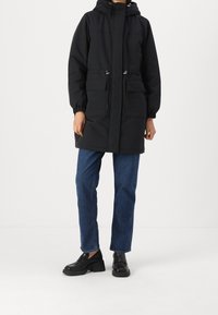 Parka nera imbottita con cappuccio, polsini elasticizzati, due tasche frontali e vita con cordino. Indossata con jeans blu a gamba dritta e scarpe nere.
