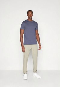 Homem em pé contra um fundo branco, vestindo uma t-shirt azul ajustada, calças de jogging bege e tênis brancos, de frente com uma postura relaxada.