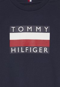 Granatowy bawełniany sweter z okrągłym dekoltem z prostokątnym logo w kolorze białym, czerwonym i niebieskim z wyrazami "TOMMY" i "HILFIGER" napisanymi dużymi literami.