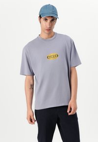 Lys grå oversized t-skjorte med en sirkulær gul "GUESS" logo på brystet. Myk bomullsduk. Båret med en blå denim cap.