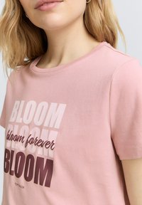 Camiseta de algodón rosa con tipografía en negrita que dice "BLOOM" en varios tonos, cuello redondo y mangas cortas. Ajuste casual.