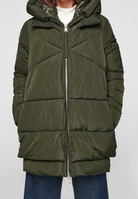 Chaqueta de plumas verde oliva con un acabado brillante, que cuenta con capucha, cierre frontal con cremallera y un patrón de acolchado en forma de Chevron, combinada con jeans de corte relajado.