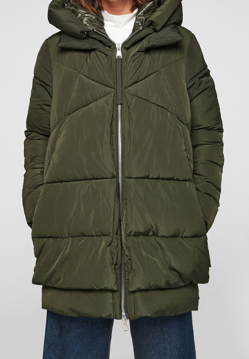 Chaqueta de plumas verde oliva con un acabado brillante, que cuenta con capucha, cierre frontal con cremallera y un patrón de acolchado en forma de Chevron, combinada con jeans de corte relajado.