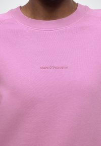 Rozā džemperis ar ribotu apaļu kakla izgriezumu. Uz krūtīm ir smalks, reljefs "Marc O'Polo DENIM" uzraksts. Gluda tekstūra.