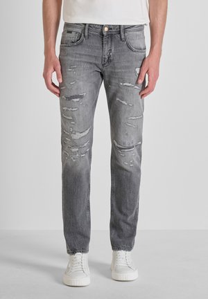 Man draagt nauwsluitende grijze versleten spijkerbroek en witte sneakers, staand tegen een effen lichte achtergrond.