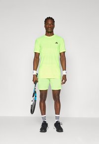 Tenue de sport neon verte avec manches courtes et shorts, ornée de rayures texturées. Chaussettes noires et blanches et baskets, tenant une raquette de tennis.
