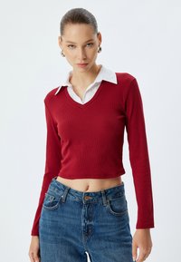 Punainen ribbipinta pitkähihaine crop-top, jossa on V-pääntie, valkoisen kauluspaidan päällä. Yhdistetty korkeavyötäröisten sinisten farkkujen kanssa.