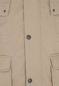 Beige bomuldsjakke med to brystlommer, central knaplukning og sølvfarvede metaldele. Glat og robust tekstur.