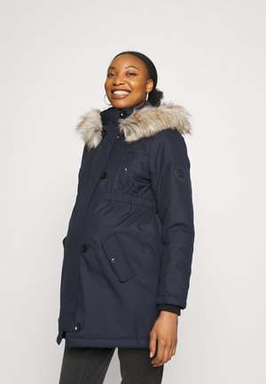 Parka marine avec capuche en fausse fourrure, taille élastique, poches avant et fermeture à bouton-pression, présentant un tissu lisse et texturé.