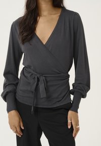 Graues Wickeloberteil mit langen, gepufften Ärmeln und einer Schleife in der Taille. Weicher Stoff mit tiefem V-Ausschnitt und gerippten Bündchen. Kombiniert mit schwarzen Hosen.