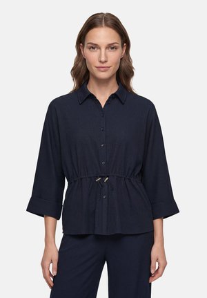 Femme aux cheveux bruns portant un chemisier bleu marine boutonné avec des manches trois-quarts et une taille à cordon, debout devant un fond uni.