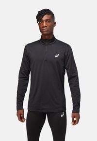 Black long-sleeve athletic shirt with a quarter-length zip, featuring a reflective logo on the chest and a smooth, dry-fit material.