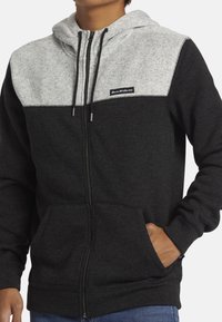 Hoodie zippé noir et gris avec un panneau supérieur gris clair, poches kangourou, capuche avec cordon de serrage et patch logo sur la poitrine.
