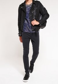 Man som går iförd svart läderjacka, grafisk t-shirt, slim fit svarta byxor, svarta slip-on skor och bär en svart läderryggsäck.