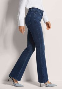 Flared dark blue denim jeans with a high waist, pair of back pockets, and a smooth texture, worn with pointed light blue heels.