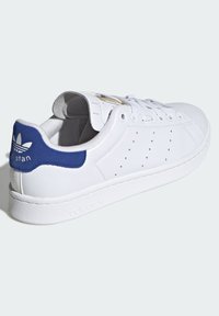 Bílé kožené tenisky s modrou semišovou patou, perforovanými detaily po stranách, plochou bílou gumovou podrážkou a zlatými logovými akcenty.