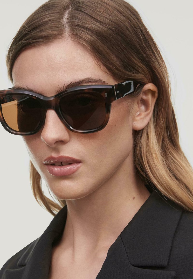 Lunettes de soleil écaille de tortue marron avec des verres sombres, de conception angulaire surdimensionnée, et des accents noirs sur les branches, comportant des détails de marque subtils.