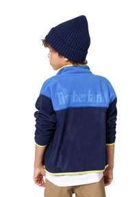 Veste en polaire bleu marine avec une section supérieure bleu clair et des accents jaunes. Présente un logo dans le dos et des poignets côtelés. Portée avec un bonnet assorti.