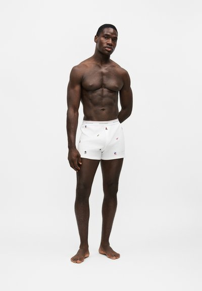 Homme musclé à la peau foncée debout pieds nus, portant un boxer blanc avec de petits motifs colorés sur un fond clair uni.
