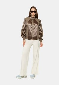 Giacca bomber in satin marrone con colletto alto, abbinata a pantaloni bianchi a gamba larga. Il modello indossa occhiali da sole scuri e scarpe da ginnastica azzurro chiaro.