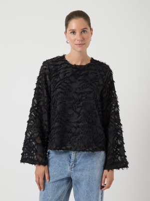 Svart teksturert topp med lange ermer, lagdelt stoff med frynsete detaljer og rund hals, kombinert med lyseblå jeans.