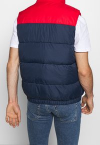 Prošívaná vesta s červenou horní částí a tmavě modrým tělem. Hladká textura s elastickým pasem. Nositelná přes bílou košili a modré džíny.