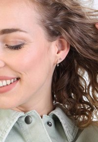 Silberne Ohrhänger mit einem herabhängenden Kristallakzent. Das Model hat lockiges Haar, trägt eine mintgrüne Jacke und lächelt leicht. Dezentes Make-up.