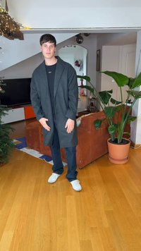 Długi ciemnoszary płaszcz z kołnierzem, noszony na czarnej koszuli i ciemnych spodniach. Białe trampki, stojący na drewnianej podłodze, w tle rośliny.