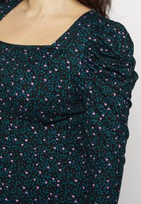 Robe noire avec un décolleté carré, ornée de motifs de cœurs turquoise et violets. Le tissu est froncé aux épaules, créant du volume.