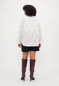 Λευκό πουκάμισο με μακριά μανίκια και κόκινες καρδιές, σε συνδυασμό με μαύρες σορτς και μπορντό μπότες μέχρι το γόνατο. Απλός, χαλαρός σχεδιασμός.