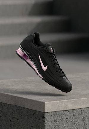 Chaussure de course athlétique noire et rose avec semelle à air visible, flottant au-dessus d'un rebord en béton sur un fond flou d'escaliers gris.